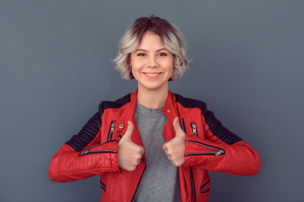 年轻女子穿着皮夹克孤立地站在灰色的墙上竖起大拇指图片下载