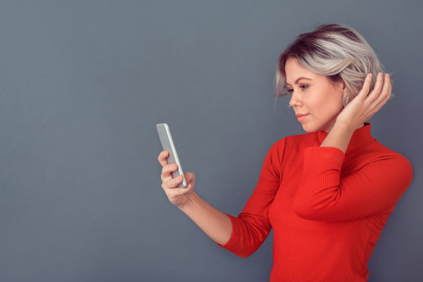 身穿红色衬衫的年轻女子手持智能手机孤立在灰色的墙上图片下载