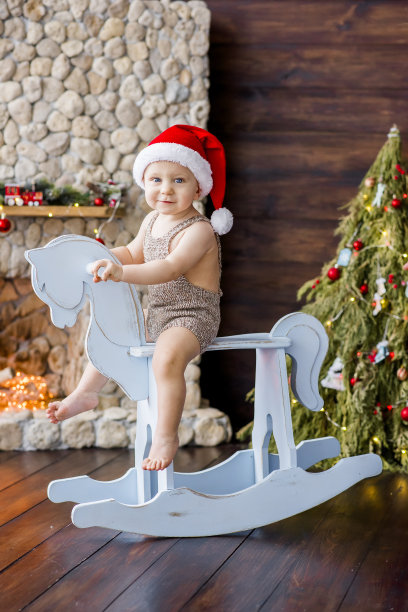 一个小男孩骑着一匹老式的木制马，旁边是一棵挂着彩灯花环的圣诞树，还有一个石头壁炉。孩子穿着针织连体衣，戴着圣诞老人帽。拷贝空间，贺年卡图片下载
