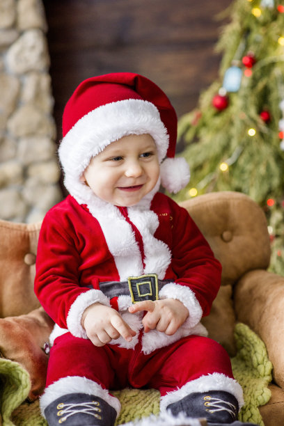 一个小男孩的特写，打扮成一个红色的圣诞老人，玩在一个石头壁炉与明亮的花环，把一个礼物盒下的圣诞树。新年贺卡。宝宝笑了，玩得很开心。图片下载