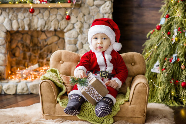 一个穿着红色圣诞老人服装的小孩，坐在家里的沙发上，手里拿着一份礼物。新年贺卡。金发男孩笑了。石头壁炉旁的圣诞树和盒子图片下载