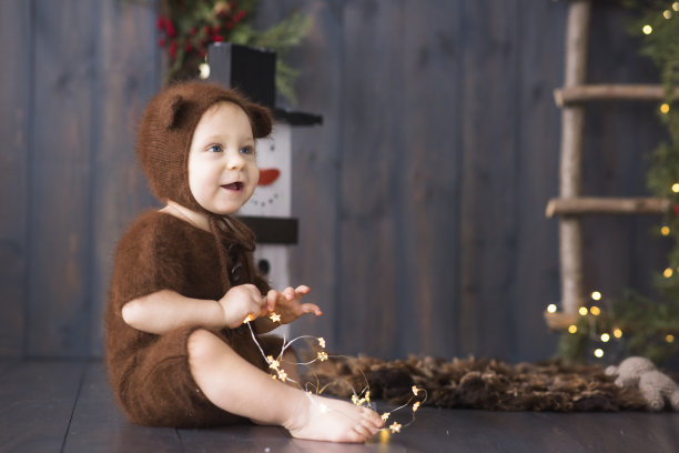 一个快乐的小男孩穿着棕色，毛茸茸的，编织的熊的服装，坐在furon的木地板上，玩着一个明亮的圣诞花环的灯。附近有一个雪人。新年贺卡。本空间图片下载