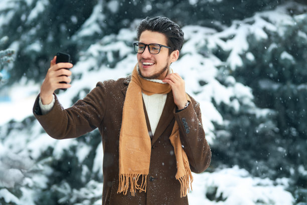 一名穿着外套、毛衣、围巾和眼镜的时尚男士在雪天公园里自拍。图片下载
