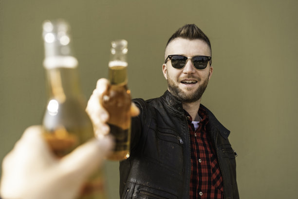 不刮胡子的酷男和最好的朋友在户外以绿色墙壁为背景敬酒-穿着皮夹克的时尚男子的手与啤酒瓶敬酒-友谊的概念，关注男人的脸图片下载