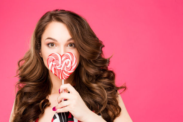 漂亮的年轻女子手里拿着糖果笑得很灿烂。时尚的女孩与明亮的妆容，在她，孤立在粉红色的背景图片下载