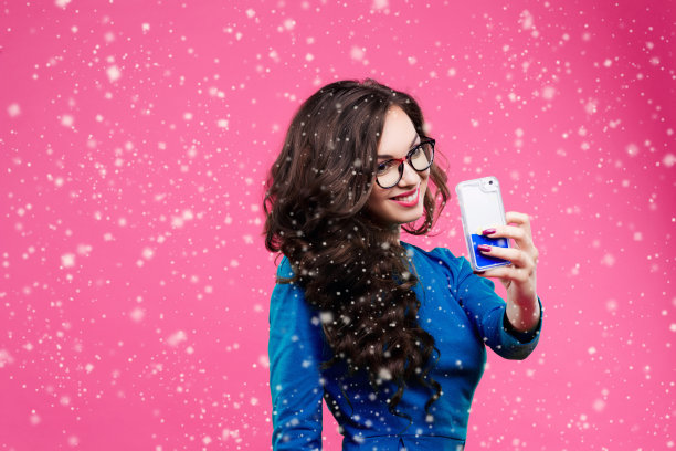 漂亮的黑发女孩与长波浪的头发突出，并在她的智能手机上自拍。穿着绿色的夹克和牛仔裤。在粉红色的工作室背景上摆姿势图片下载