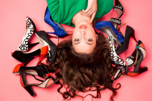 从上面看，一个漂亮的黑发女孩躺在粉红色的地板上，有着长长的卷发和蓝色的妆容，她的头周围穿着不同的时尚鞋。手摸下巴，看镜头图片下载