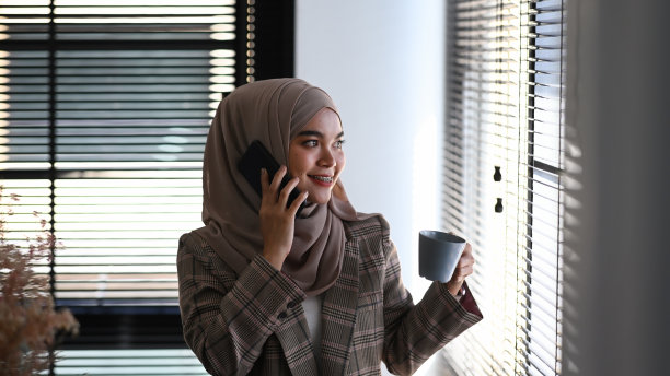 一位戴着头巾的穆斯林妇女站在办公室里一边打电话一边拿着热饮。图片下载