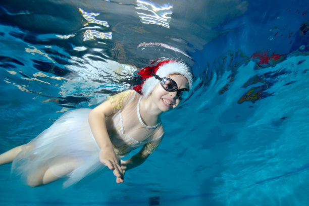 一个面带微笑的小女孩在儿童游泳池游泳，在镜头前摆姿势，她戴着圣诞老人的帽子，穿着白色的裙子，蓝色的背景。的概念。水平方向图片下载