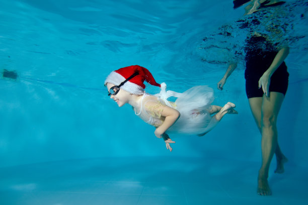 可爱的小女孩正在一个教练的指导下在儿童游泳池里潜水。她戴着圣诞老人的帽子，穿着白色的裙子，微笑着在水下游泳。肖像。图像的水平方向图片下载