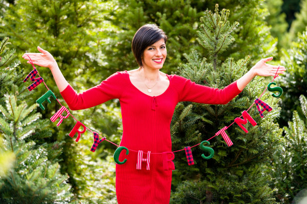 红色连衣裙上微笑的女人举着“圣诞快乐”横幅在圣诞树农场。图片下载