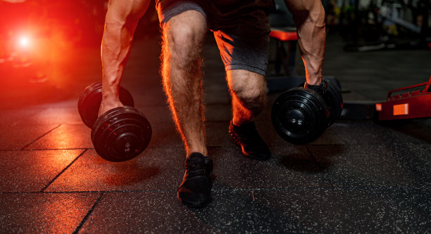 一个强壮的运动员从地板上拿哑铃的剪裁照片。在现代化的健身房锻炼。温暖的橙色灯光。图片下载