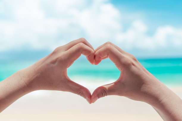 女人的手在蓝天和海滩的背景上做心形。图片下载
