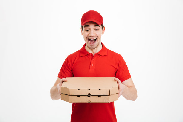 照片中，一名身穿红色t恤、头戴红色帽子的快递员拿着两个披萨盒子在白色背景下给外卖人员点餐图片下载