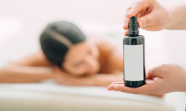 芳香按摩广告。特写按摩油在一个按摩治疗师的手中，在背景，一个女人按摩后。图片下载