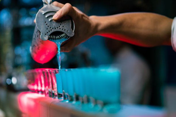 专业调酒师的手从鸡尾酒调酒器将混合后的蓝色烈性酒倒入吧台的小酒杯中，为夜总会的顾客提供服务。男调酒师在酒吧为顾客准备酒精饮料图片下载