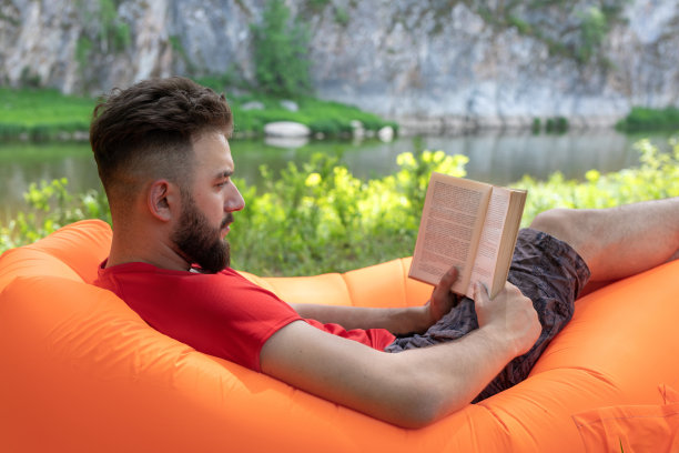 长胡子的男人或男人正躺在户外的橙色充气躺椅或沙发上看书图片下载