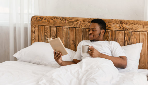 千禧一代黑人男人躺在床上看书喝咖啡图片下载