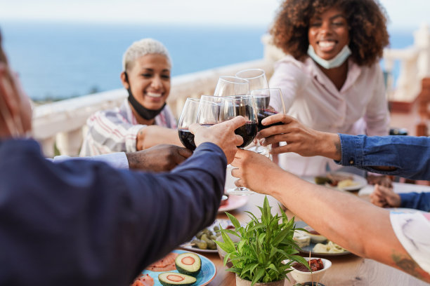 快乐的多种族人士在家里的露台上享受晚餐，并在下巴下戴着外科口罩时用红酒欢呼图片下载