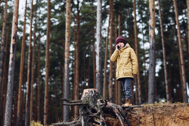 一个穿着黄色夹克的男孩正站在树林里打电话。图片下载