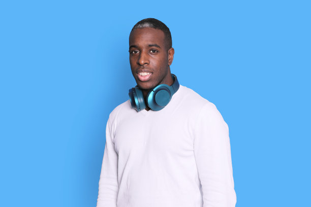 英俊的黑皮肤男子戴着耳机在他的脖子上，看着相机在蓝色的背景图片下载