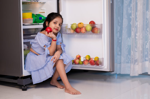 可爱的小女孩手里拿着水果，坐在冰箱里。图片下载