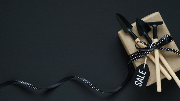 黑色星期五的网上购物。棕色礼盒，花园工具与黑色蝴蝶结在白色的豌豆和黑色的标签在黑色的背景，俯视图，平面lay，复制空间。园林工具销售理念。图片下载