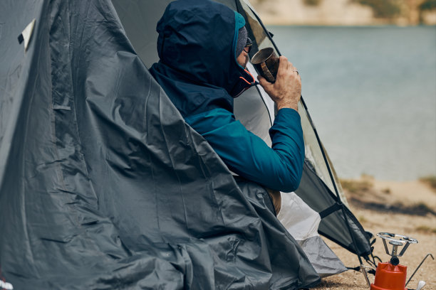 一位探险家坐在湖岸的帐篷里，看着水，喝着咖啡。图片下载