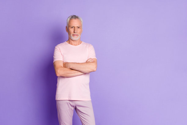 照片中自信的老人面无表情，双手合十，穿着粉红色的裤子，t恤孤立的紫色背景图片下载