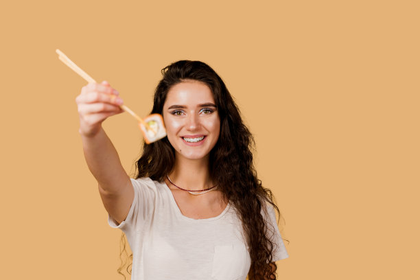 快乐的女孩把寿司送给孤立在黄色背景上的你。年轻的女孩拿着筷子寿司，微笑，有乐趣，想要吃一片卷费城。寿司外卖广告。图片下载