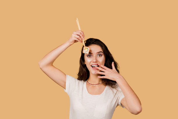 快乐的女孩与寿司隔绝在黄色的背景上。年轻的女孩拿着筷子寿司，微笑着，有乐趣，想要吃一片卷费城。寿司外卖广告。图片下载