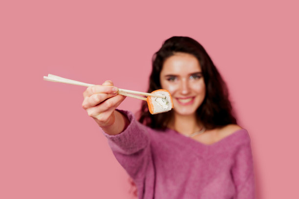 快乐的女孩给你寿司孤立在粉红色的背景。年轻的女孩拿着筷子寿司，微笑着，有乐趣，想要吃一片卷费城。寿司外卖广告。图片下载