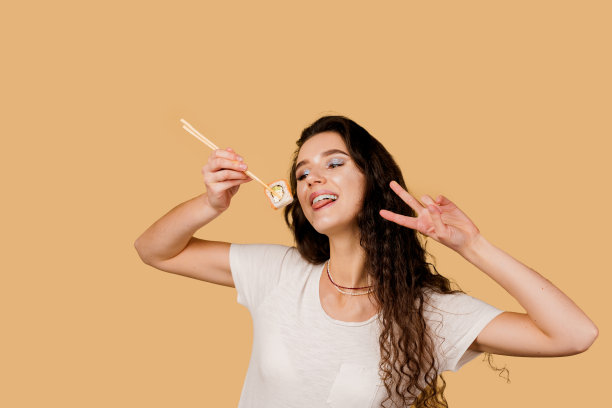 快乐的女孩与寿司隔绝在黄色的背景上。年轻的女孩拿着筷子寿司，微笑着，有乐趣，想要吃一片卷费城。寿司外卖广告。图片下载