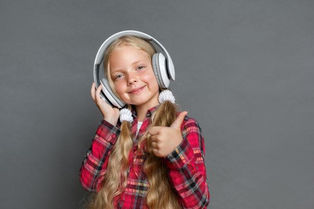 小女孩戴着耳机，扎着马尾辫，孤立地站在灰色的舞台上听着音乐，大拇指向上看着相机，令人愉快图片下载