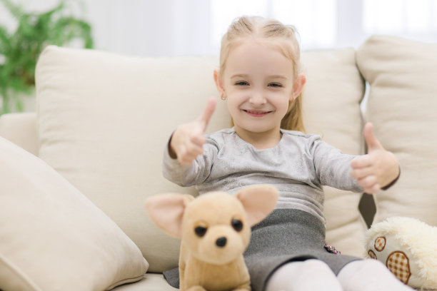 漂亮的小女孩和泰迪熊坐在沙发上，翘起大拇指。图片下载