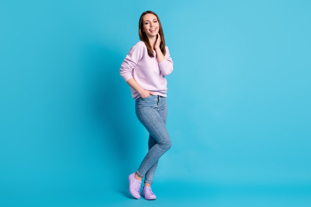 全身照片漂亮迷人的女士好心情触摸脖子梦想家喜气洋洋的微笑街道看穿休闲的紫色运动衫套头衫鞋子牛仔裤孤立的蓝色背景图片下载