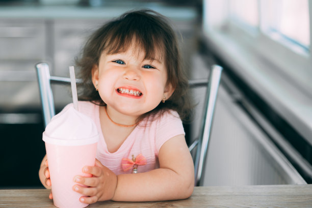 一个穿着粉红色裙子、蓝眼睛的小女孩坐在厨房里，拿着一只粉红色的鸡尾酒杯喝着酒，做着鬼脸图片下载