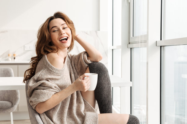 愉快的年轻女子在客厅享受消遣的照片，坐在靠近窗户的扶手椅上，喝一杯茶或咖啡图片下载