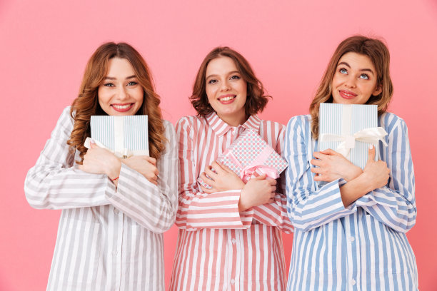 照片上的女孩，20多岁的朋友，穿着家装，拿着礼盒，在睡衣派对上表达兴奋，在粉红色的背景下孤立图片下载