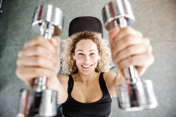 背景上，一个女人正躺在一个训练器械上。她的手拿着两个哑铃在前台。图片下载