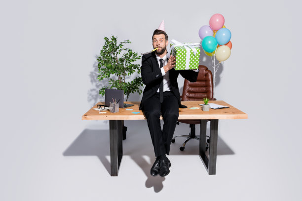 英俊的商人的全身照片坐在桌子上拿着大礼盒吹噪音的人遇到同事办公室惊喜生日派对穿服装套装孤立的灰色背景图片下载