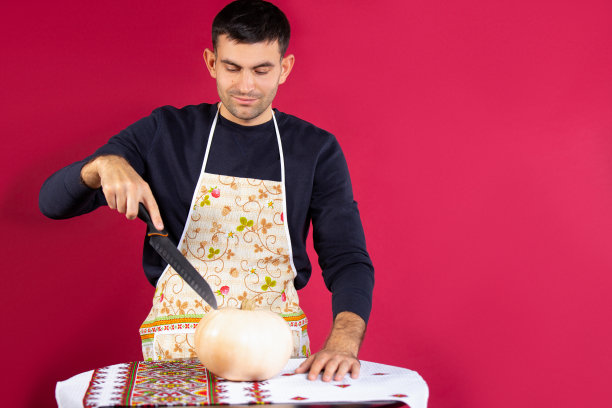 一个穿着厨房围裙的男人用刀切南瓜。照片的粉红色背景与空白。反对性别陈规定型观念。图片下载