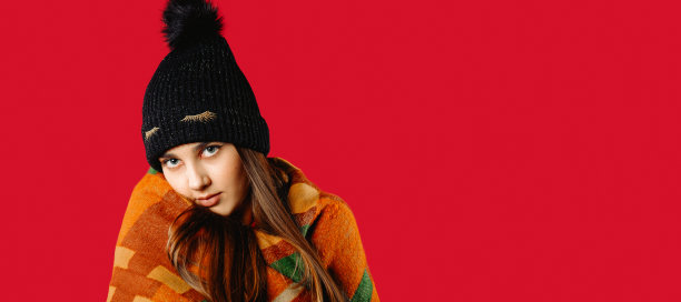 横幅,长格式。一个可爱的女孩在一个冬天的帽子和包裹在一个温暖的格子，摆姿势在一个红色的照片与侧空间。冬天的概念图片下载