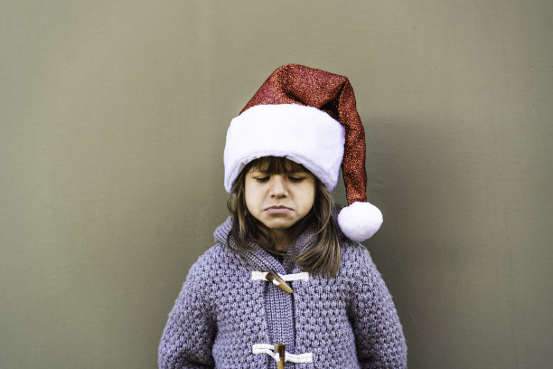 悲伤的小女孩穿着圣诞老人帽子对着绿色的墙壁背景在圣诞时间户外-小女孩生闷气不想要的圣诞礼物-不快乐的童年概念圣诞节假期图片下载