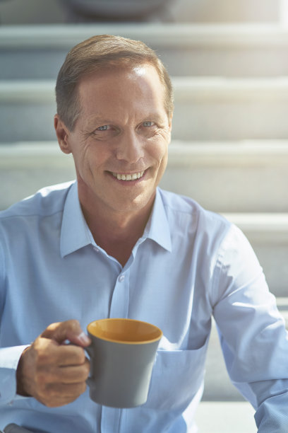 快乐成熟的商人在衬衫拿着一杯咖啡，看着相机和微笑，在办公室休息，垂直拍摄图片下载