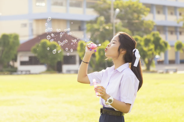 亚洲可爱的女孩穿着白色校服在草坪上玩泡泡气球。可爱的女孩在花园里玩肥皂泡。图片下载