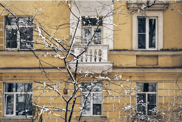 带有白色阳台的旧建筑的正面。房子前面的树被雪覆盖着。明信片上有一个安静的城市冬季老庭院。图片下载