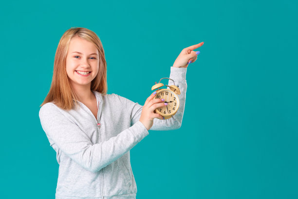 一个十几岁的女孩拿着一个闹钟，站在一个孤立的绿松石背景上，开心地笑着。在你的文本空白区域做一个手势图片下载