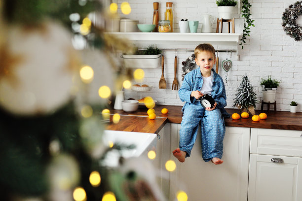 一个穿着蓝色睡衣的小男孩光着脚坐在一张木桌上，背景是现代风格的白色厨房和挂在圣诞树上的圣诞彩灯，上面挂着一个闹钟。图片下载