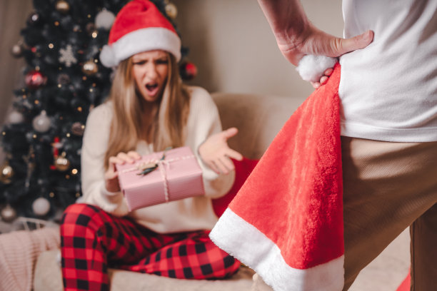 近距离后方的愤怒的男人拿着圣诞帽和不快乐的年轻女子与圣诞礼物盒靠近圣诞树。几个问题的概念图片下载